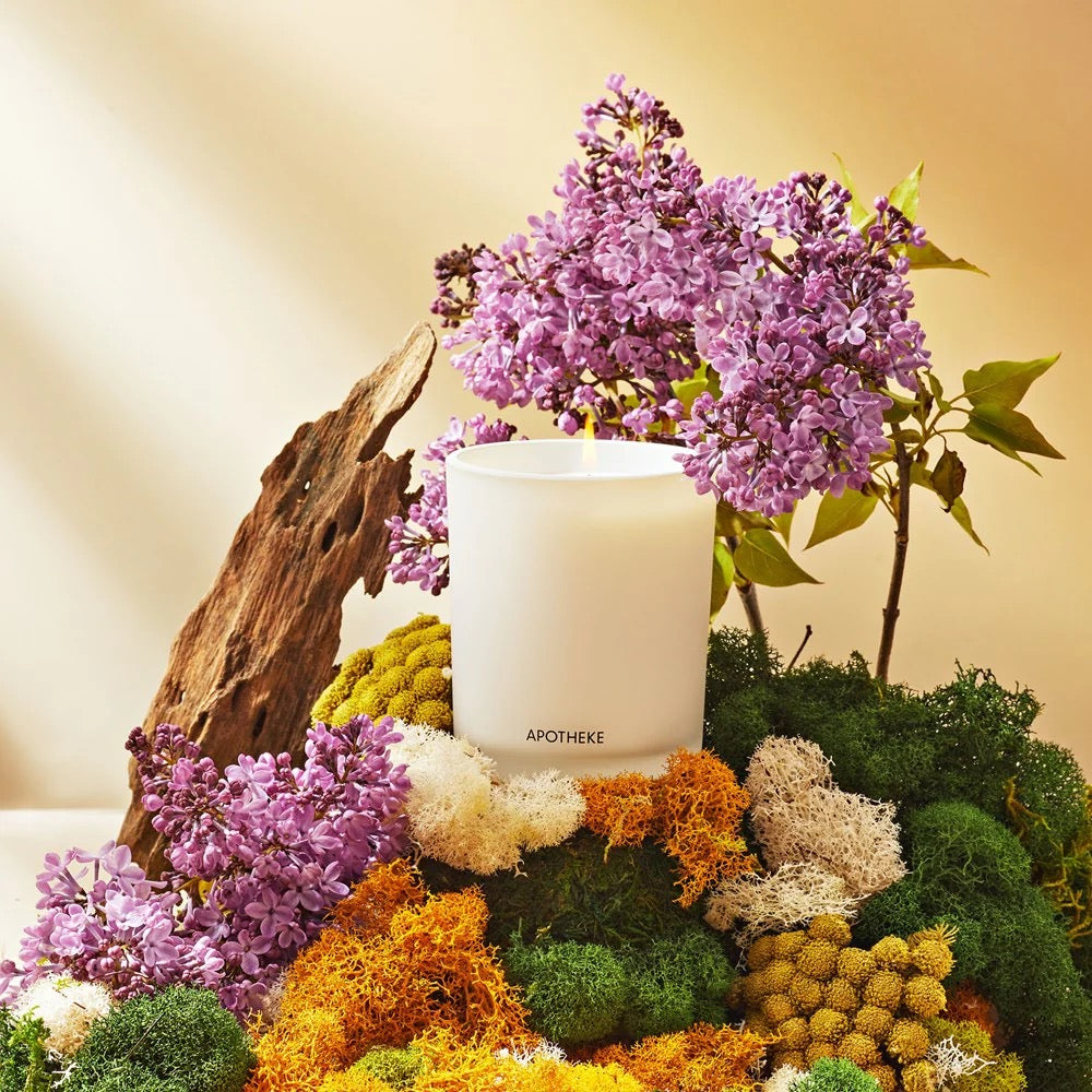Apotheke signature candle in a minimalist white glass jar surrounded by blooming lilacs, colorful moss, and natural wood—an elegant floral-scented home fragrance perfect for spring styling, thoughtful gifting, and everyday luxury from Willow Boutique.