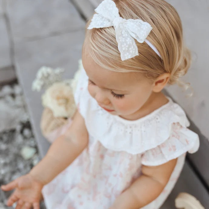 Baby girl wearing a soft floral dress with eyelet ruffle collar and a white lace bow headband, sitting outdoors—perfect for special occasions, spring family photos, or everyday heirloom-inspired baby fashion from Willow Boutique.